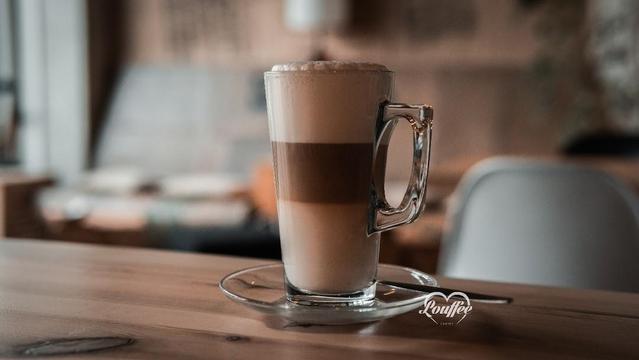 A glass of latte artfully layered on a saucer with a spoon, sitting on a wooden table.