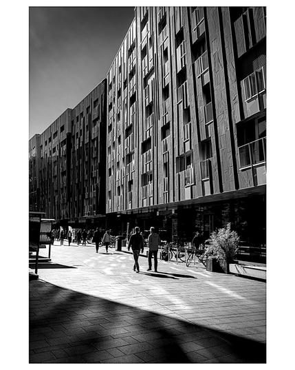 A high-contrast black and white street scene features a long, modern building with a segmented, vertical-panel facade and small windows dominating the upper half. Below, a wide, paved pedestrian walkway is sharply divided by a dramatic, diagonal shadow that cuts across the lower left. Several figures walk along the sunny sidewalk toward the background. The strong midday light emphasizes the building's texture and creates deep, geometric shadows on the pavement. The scene captures a lively urban street with striking modern architecture.