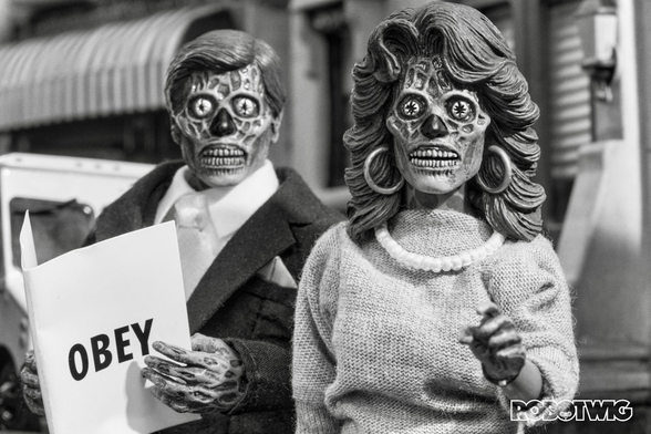 Two Aliens dressed in Hunan clothes look at you one holds a newspaper with the word Obey written on it