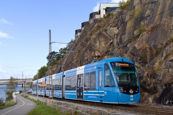 Une rame de tram de couleur bleue circule sur une voie ferrée coincée entre un chemin étroit goudronné et un relief rocheux. En haut de ce relief, on voit des maisons. Le chemin se trouve le long d’une côte ; on voit l’eau et un pont en arrière-plan.

La rame est de type CAF Urbos dans la livrée des transports de Stockholm. Elle porte le numéro 559. La girouette affiche « 21 Gåshaga Brygga ».