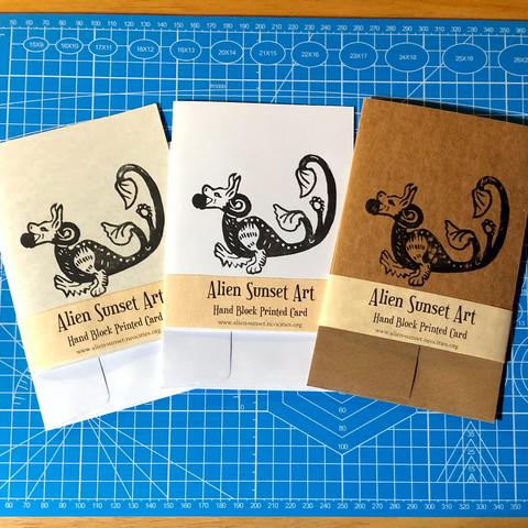 a set of three cards laid out on a blue cutting mat. a brown Kraft card, a plain white card, and a cream parchent pattered card. each card has a yellow parchemnt strip of paper wrapped around it that says "Alien Sunset Art" Hand Block Printed Card.

the block print on the cards is of a small lizard like creature with two furry legs, a long split tail with leaved at the end, and a dog's head carrying a ball in it's mouth