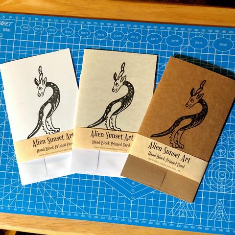 a set of three cards laid out on a blue cutting mat. a brown Kraft card, a plain white card, and a cream parchent pattered card. each card has a yellow parchemnt strip of paper wrapped around it that says "Alien Sunset Art" Hand Block Printed Card.

the block print on the cards is of a tall two legged creature with a long tail, furry legs and a horses head and unicorn horn