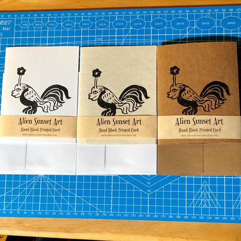 a set of three cards laid out on a blue cutting mat. a brown Kraft card, a plain white card, and a cream parchent pattered card. each card has a yellow parchemnt strip of paper wrapped around it that says "Alien Sunset Art" Hand Block Printed Card.

the block print on the cards is of a small creature with two paws, a rooster's tail, small wings and a face like a smug bulldog with a flower growing out of it's head
