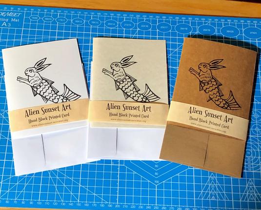 a set of three cards laid out on a blue cutting mat. a brown Kraft card, a plain white card, and a cream parchent pattered card. each card has a yellow parchemnt strip of paper wrapped around it that says "Alien Sunset Art" Hand Block Printed Card.

the block print on the cards is of a swimming merbunny, - a bunny with a fish tail
