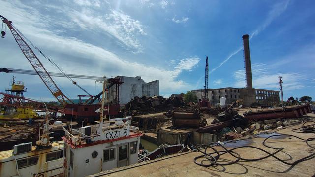 Foto Panorámica de desguazadero naval, detrás de las pilas de hierros oxidados se ve lo que alguna vez fue un edificio industrial abandonado. Adornan el paisaje grúas y lingas de acero, así como algunos barcos atracados
