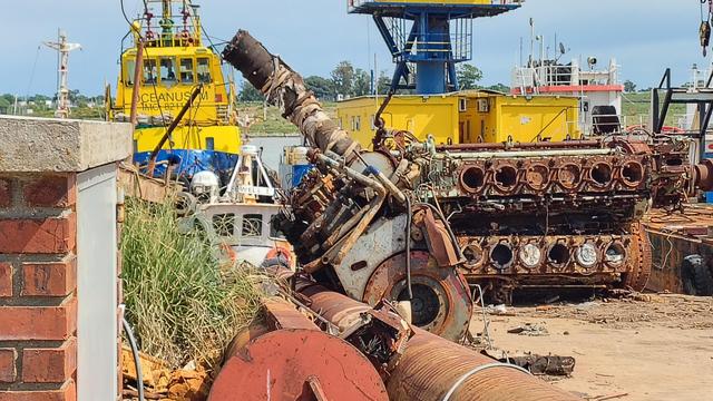 Foto de desguazadero naval donde se ve un motor de navío desarmado, el.bloque de  12 cilindros supera en tamaño a una camioneta. En el fondo se ven dos varcos remolcadores aamarillos y azules