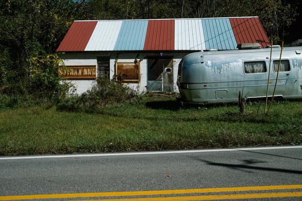 A photo of a vintage Airstream-style trailer parked in a grassy area in front of a small, single-story cinder block building that used to be a roadside market store. The building has a striped red, white, and blue metal roof and is surrounded by overgrown vegetation, with a line of trees in the background.