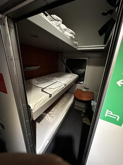 A compact sleeper cabin with two stacked beds, clean white bedding, and a small table. The interior features a wooden panel, a window, and some personal items on the floor. A green sign indicates a seating option.