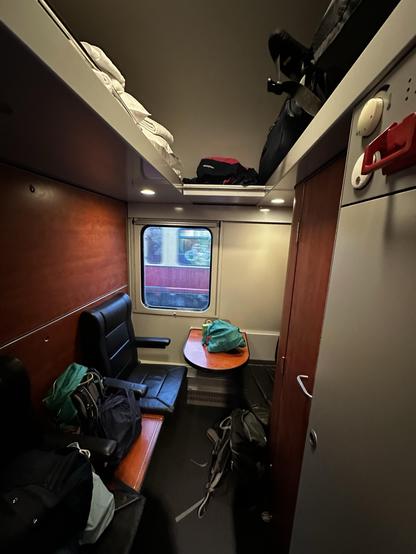 An interior view of a sleeper compartment on a train, featuring two black leather seats, a small wooden table, and overhead luggage storage with bags. Clean towels are stacked above, and there are several bags on the floor. A window shows a glimpse of a blurred train.