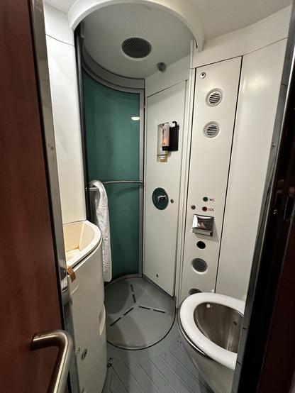 A compact bathroom featuring a toilet, curved glass door, a small sink with a tap, and buttons on the wall for functions like "WC" and "SOS." The interior is primarily white with some teal accents.
