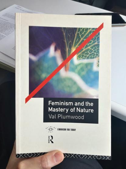 A hand holds a copy of the book " Feminism and the Mastery of Nature" by Val Plumwood.