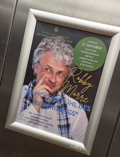 Ein Plakat in einem silbernen Rahmen kündigt eine Veranstaltung an. Es zeigt einen Mann mit grauen Locken, Brille und kariertem Schal. Der Text informiert über einen Kabarett-Abend mit Robby Mörre unter dem Titel „Im Hochgefühl des Niedergangs“, der am Freitag, 17. Oktober um 20 Uhr stattfindet.