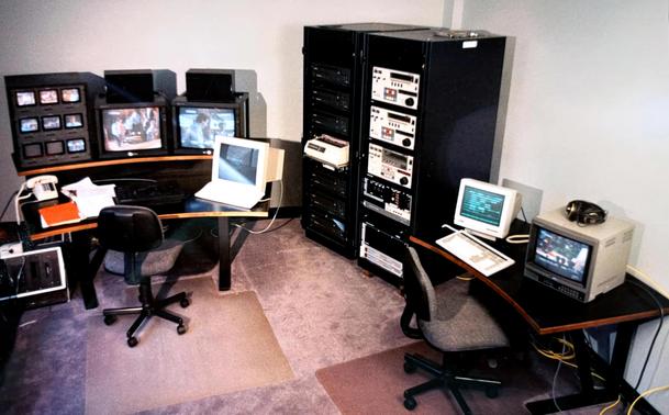 A vintage multimedia control room featuring several monitors displaying video footage, a couple of computers, and audio/video equipment organized in a rack.