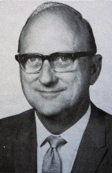 A black-and-white portrait of a softly-smiling C. Sumner Wemp. He is a middle-aged white fellow with hair thinning on top. He wears browline eyeglasses and coat-and-tie.