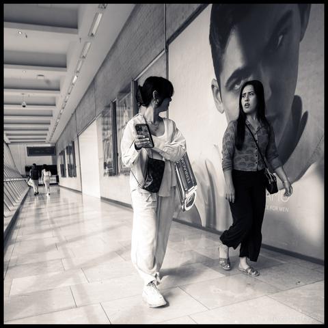 Black and white photograph of two women walking through a modern shopping corridor in Birmingham, England, one looking at her phone wearing light-coloured clothing with shopping bags, the other in darker attire standing beside a large fashion advertisement showing a male model's face.
