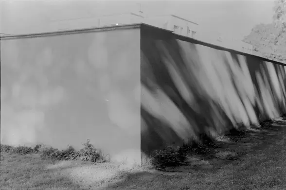 ein schwarzweißes bild, stark überbelichtet. eine mauerecke, links fast weiß, rechts zeichnen sich schatten ab. im hintergrund sind gebäude angedeutet.