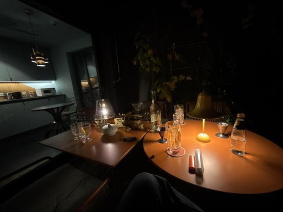 weitwinkel-aufnahme eines runden und eines quadratischen tisches auf der dachterasse des lyf east-hotels im frankfurter ostend; warm beleuchtet - noch ein gast am tisch und im hintergrund die community-kitchen im 6. stock