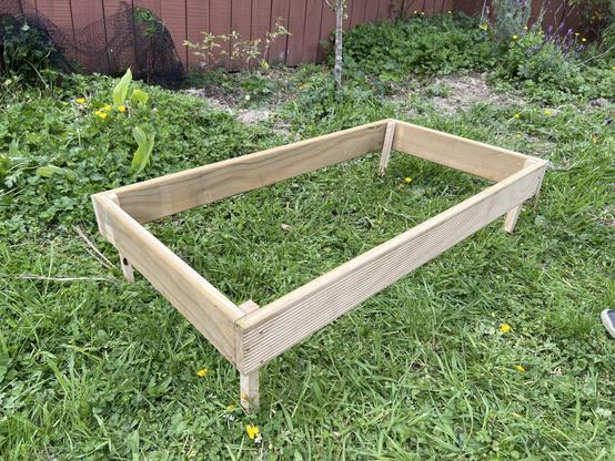 A wooden garden bed frame is positioned on grass, showcasing its rectangular shape and elevated spike legs, not yet in the ground. The background features green foliage and a wooden fence.