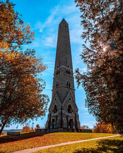A very tall obelisk that has an ornate Gothic at the bottom third of the structure is at the center of the image. The structure is framed by two yellow-red-green trees on both sides, and there are fall leaves on the ground. The sky behind the obelisk is a light blue with not many clouds in it.