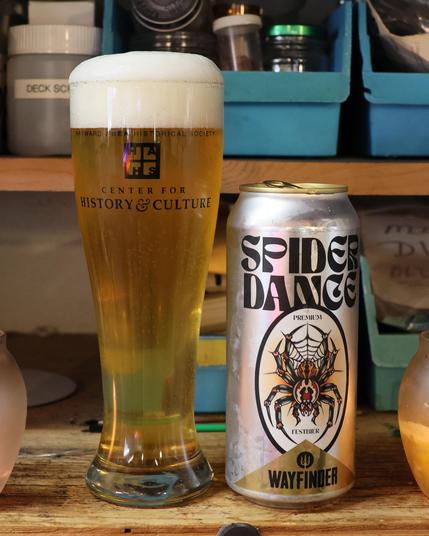 A pilsner glass filled with a light amber liquid stands next to a beer can. The can is silver, with a drawing of a large black tarantula on the label. The label reads “Spider Dance Premium Festbier, Wayfinder.”
