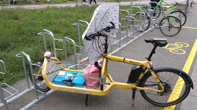 A yellow Bullitt cargo bike with an open cargo bay with just a few things tied to it, has a foldable white umbrella with many dots in a few bright colors mounted vertically in front of the handlebar with the umbrella's handle pointing towards the saddle.