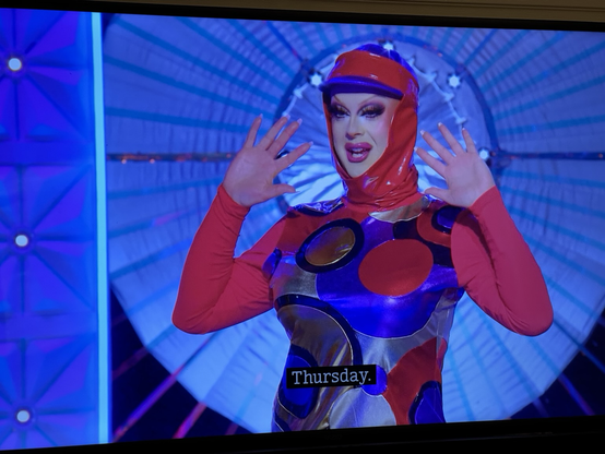 On the Drag Race UK runway, Bonnie Ann Clyde, dressed as Patsy from AbFab from one of the sixties flashback moments in the red and purple outfit, holds her hands up on either side of her face and says “…Thursday.”