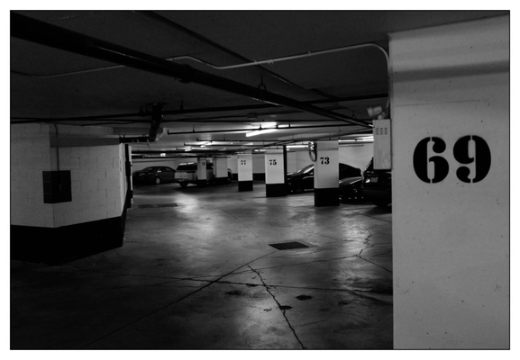 Black and white photograph of an underground parking lot with multiple cars parked. A pillar in the foreground prominently displays the number 69. The environment is dimly lit with overhead lights and includes visible numbering on other pillars, guiding to different parking spots.
