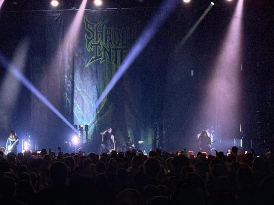 Shadow of Intent performs on stage with colorful lights shining, and a large audience watching the concert.