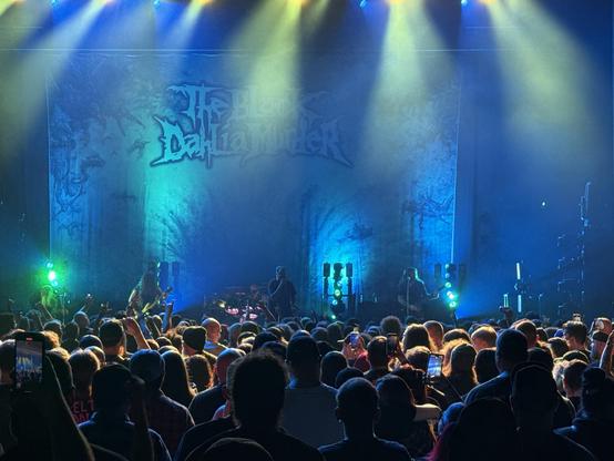 A live concert with The Black Dahlia Murder performing on stage, colorful lights, and a large audience watching and recording.
