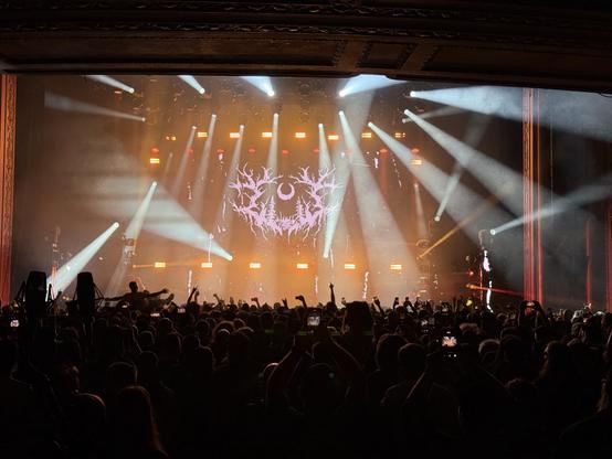 Lorna Shore with bright stage lights, a crowd of fans, and a dark backdrop featuring a stylized logo with a crescent moon and tree branches.