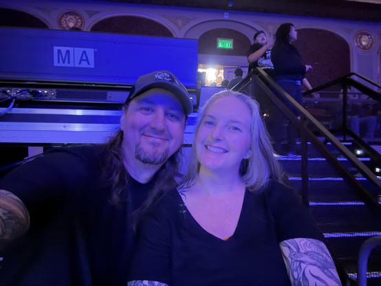 A man with long hair and a cap, and a woman with blonde hair smiling, taking a selfie inside a dimly lit venue with stairs and people in the background.