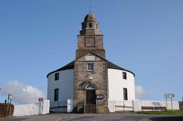 The Round Church in Bowmore. The image shows a view up a road that is glimpsed in the bottom of the frame to a two-storey circular church. Much of the structure is white, though the front entrance, which is facing us, is made of stone and is topped off by a tower and bellcote. The front door is accessed by stone steps. The sky is largely blue.