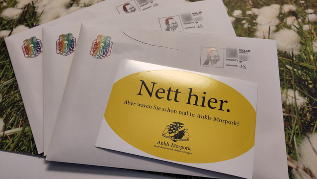 Drei Umschläge liegen überlappend auf dem Tisch. Auf jedem Umschlag in der linken oberen Ecke das Logo der Wache von Ankh-Morpork in Pride-Farben. Rechte obere Ecke eine Briefmarke mit der charakteristischen Karikatur, die FlohEinstein darstellt, und eine verschwommene Webstamp-Briefmarke. Unter allem liegt eine Schreibtischauflage mit einem Bild von schmalblättrigem isländischem Wollgras.
Die Adresse des Empfängers wird von einem gelben Sticker verdeckt. Darauf steht in schwarzer Schrift "Nett hier. Aber waren Sie schon mal in Ankh-Morpork? Stadt der tausend Überraschungen" im Stil der Nett hier-Kampagne von Baden-Württemberg