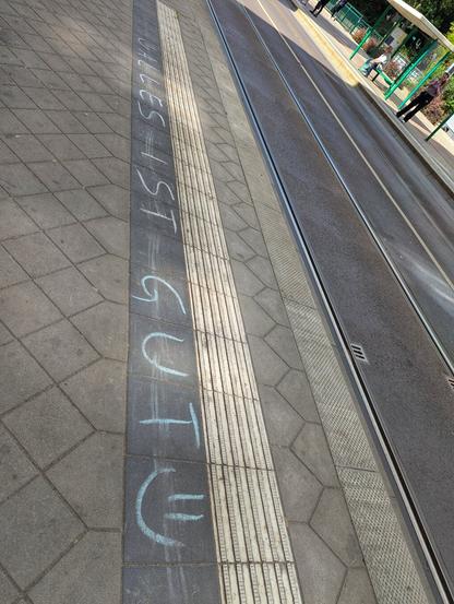 A close-up photograph of a tram platform edge at an outdoor station. The platform features tactile paving strips designed to assist visually impaired individuals. Chalk writing on the platform surface reads 'ALLES IST GUT' with a small smiley face drawn next to the text. The platform is constructed of grey, interlocking paving stones, and the tram tracks run parallel to the platform edge. In the background, there are green railings and some greenery, indicating an urban outdoor environment.