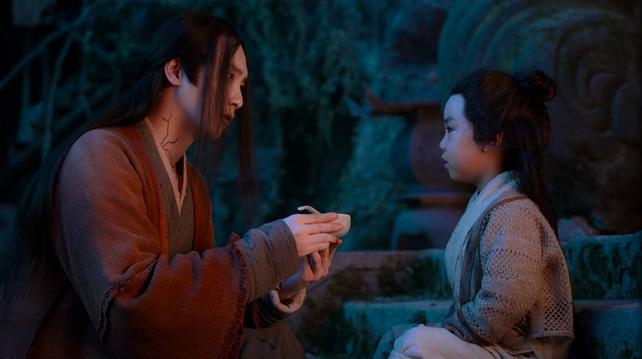 In the Burial Mounds at evening, Wen Ning kneels in front of A-Yuan, seated on a stone step, and holds up a bowl which has the handle of a utensil protruding above the top edge.