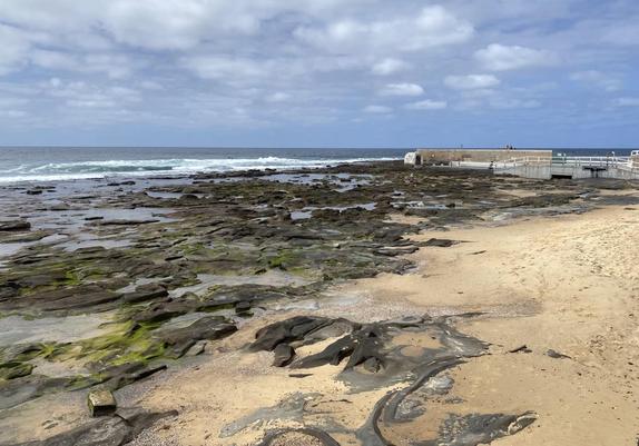 Looking southwards over the Cowrie Hole beach towards the Ocean Baths and the ocean horizon. It is low tide, exposing more of the small sandy beach and the natural rocky platform that stretches out about 20 metres before the white line of breaking waves is visible.