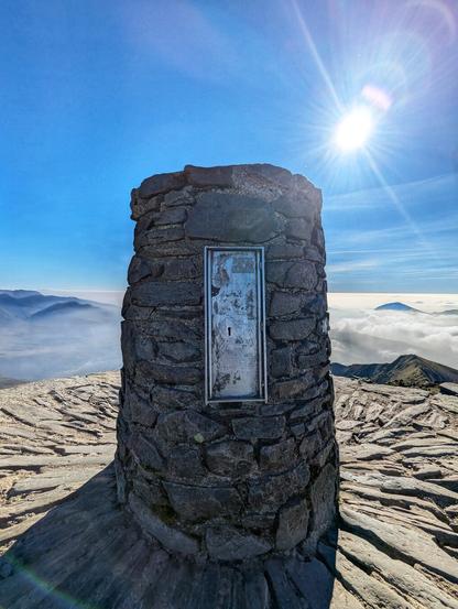 A stone summit cairn, constructed from layered rocks, stands prominently on a mountain peak. The pillar features a small metal plaque on its front. The sun shines brightly in the clear blue sky above, casting a radiant glow and lens flare around the pillar. Below, a breathtaking view of rolling hills and a sea of clouds stretches into the distance. On the right side of the pillar, there is a small black graffiti drawing of a cat peeking around the corner. The scene captures a serene and expansive landscape, typical of a mountain summit.