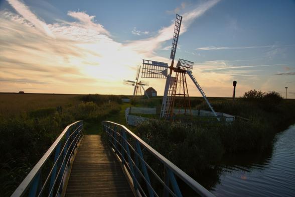 Eine alte Mühle in einem Feld neben einem Bach. Davor ist eine Brücke.
