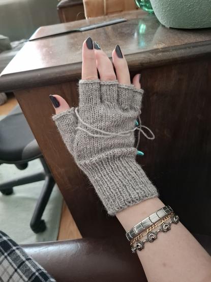 Käsi, jossa neulottu vasleanharmaa kynsikäs, langanpäät päättelemättä ja silmukkamerkkejä kiinni työssä.