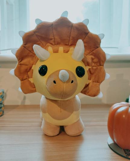 A yellow and brown triceratops plushie sat on a wooden shelf. A pumpkin candle can just be seen to its right.
