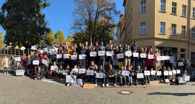 Das Foto zeigt eine große Gruppe von Menschen, die auf einer breiten Treppe im Freien stehen und sitzen.
Sie halten Blätter mit einzelnen Buchstaben hoch, die zusammen den Satz „WIR SIND DAS STADTBILD“ ergeben.
Die Gruppe besteht aus Menschen unterschiedlichen Alters, die bei sonnigem Wetter gemeinsam an einer Aktion teilnehmen.
Im Hintergrund sind herbstlich gefärbte Bäume und ein mehrstöckiges, hellgelbes Gebäude zu sehen.
Einige Personen halten bunte Schilder und Plakate, auf denen zusätzliche Botschaften stehen.