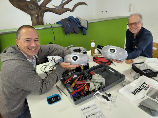 2 Reparateure halten fröhlich 2 identische Radio-CD-Player hoch, auf dem Tisch befinden sich Werkzeuge