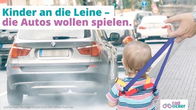 Ein Kleinkind steht an einer vielbefahrenen Straße. Es wird an einer Kinderleine von einem Erwachsen geführt. Text: Kinder an die Leine - die Autos wollen spielen.