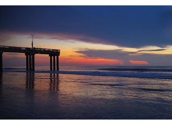 Pier silhouetted against a vibrant sunrise sky with deep orange, pink, and blue hues, reflecting on the rippling ocean waves below.