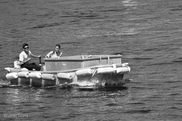 This is a black and white photo in landscape format of a garbage scow with a young man at the helm and a young woman sitting beside him. Santa Catalina (2015).
Filling most of the image is an expanse of sea water, with small ripples breaking the surface. From the centre and to the left is a small, flat bottomed work boat, about four metres long, with a light grey hull and surrounded by a string of fenders lashed to the side horizontally. The boat covers about half the width of the image and is moving from left to right. In the craft is a large rectangular metal skip, (dumpster) that occupies about two-thirds of the hull. Sitting in the stern is a young man of around twenty year old who has his left hand on the tiller. He has neat dark hair, wayfarer sunglasses, white t-shirt and dark trousers. He is looking at the camera. On his left is a female of the same age, with long blonde hair, looking down to her right with a slightly worried expression on her face. 
The location is Avalon Bay, Santa Catalina, California. The boat is a garbage scow, as in, collects rubbish from yachts moored in Avalon Harbour.