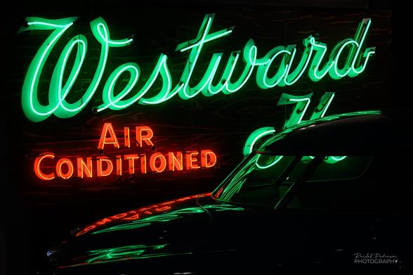 The upper rear of a 1951 Studebaker Commander Starlight Coupe reflects the light of neom signs behind it.  There is a green neon sign that says, "Westward Ho" and a read neon sign that says, "Air Conditioned".