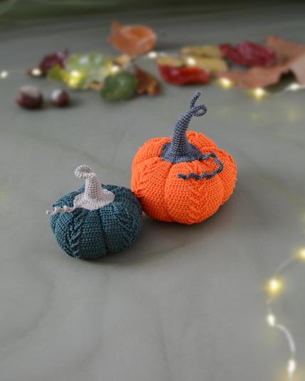 Deux citrouilles crochetées posées sur un fond vert avec une guirlande lumineuse, des feuilles d’automne et des marrons en arrière-plan : l’une est orange avec une tige gris foncé, l’autre vert foncé avec une tige grise claire et une vrille en fil assorti.