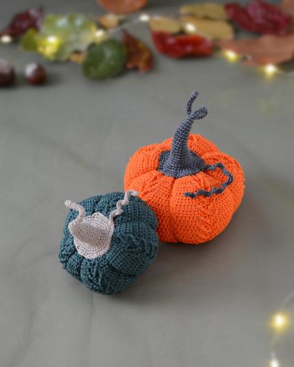Deux citrouilles crochetées posées sur un fond vert avec une guirlande lumineuse, des feuilles d’automne et des marrons en arrière-plan : l’une est orange avec une tige gris foncé, l’autre vert foncé avec une tige grise claire et une vrille en fil assorti.