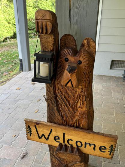 A chain-saw carved wooden bear is holding a "Welcome" sign and a lantern, while standing on a patio.  The small mantis is attached under his right arm, which is holding up the lantern.  It does look like it would tickle.