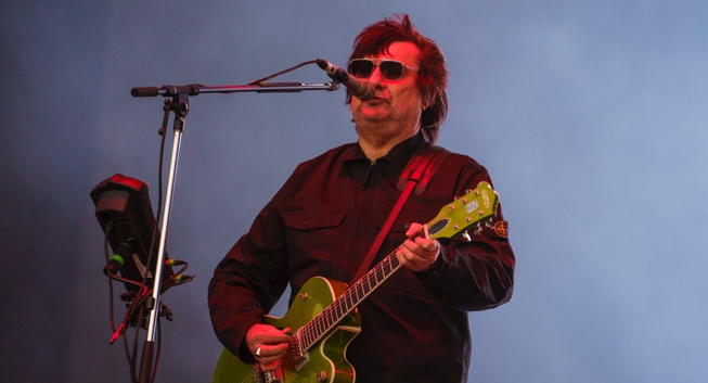 Álvaro Henríquez con gafas de sol oscuras y pelo castaño desordenado toca una guitarra eléctrica de color verde lima. Viste una camisa o chaqueta oscura de manga larga con cremallera y dos bolsillos frontales. Está de pie frente a un micrófono en lo que parece ser un escenario, con un fondo brumoso de tono azulado. A su izquierda, se ve parte de un soporte de micrófono con una pequeña unidad electrónica negra y algunos cables.